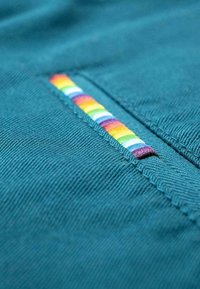 Tessuto teal con una superficie testurizzata presenta una patch a strisce orizzontali arcobaleno lungo una cucitura. Le strisce includono rosso, arancione, giallo, verde, blu e viola.