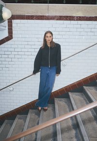 Zwarte jas met rits en oversized mouwen, gecombineerd met wijde blauwe spijkerbroeken en bruine puntige hakken, voor een betegelde trap.