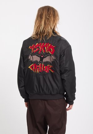 Personne aux longs cheveux de dos portant une veste noire avec une chauve-souris graphique et un texte rouge indiquant "Psycho Chiller" dans le dos.