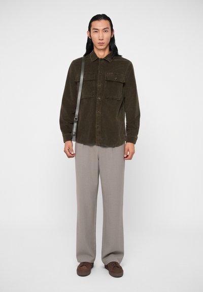 Camisa de corduroy verde escuro com dois bolsos frontais, fecho com botões, combinada com calças largas cinza claro e sapatos castanhos.