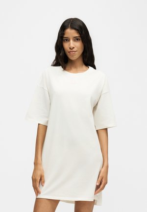Femme aux longs cheveux foncés portant une robe blanche ample à manches courtes, debout devant un fond clair et uni.