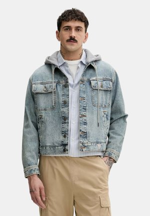 Uomo con baffi che indossa una giacca di jeans chiara con cappuccio grigio, maglietta bianca a righe e pantaloni cargo beige, in piedi con una mano in tasca.