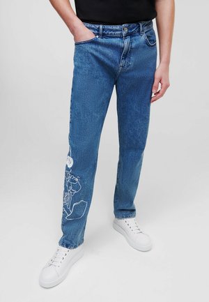 Persoon die een blauwe spijkerbroek met witte grafische ontwerpen op het linkerbeen en witte sneakers draagt, staand tegen een effen achtergrond.