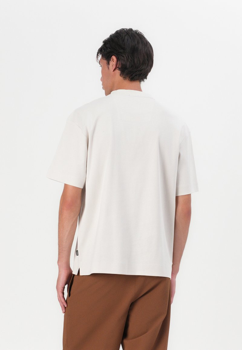 Jasno beżowa koszula z krótkim rękawem o luźnym kroju. Posiada okrągły dekolt, małe boczne rozcięcia oraz minimalną metkę z logo na dolnym brzegu.