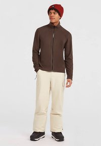 Jauns vīrietis, kurš valkā sarkanu beanie cepuri, brūnu jakas ar rāvējslēdzēju, krēmkrāsas bikses ar kabatām un melnas kurpes, stāv ar vienu roku kabatā.