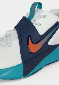 Κοντινή λήψη ενός λευκού αθλητικού Nike με γαλάζια κορδόνια, σκούρα μπλε λουρί, πορτοκαλί σήμα Swoosh και τιρκουάζ λεπτομέρειες στη σόλα και το πίσω λουράκι.