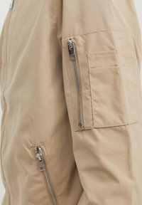Manche de veste marron clair avec deux fermetures éclair argentées verticales et une poche rectangulaire avec compartiments cousus sur le haut du bras.