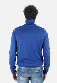 Uomo con capelli ricci che indossa un maglione blu a dolcevita e jeans azzurri slavati, di spalle verso l'osservatore.