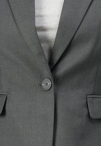 Hall kohandatud bleiser, mis on ühe korra nööbitud üle helehalli särgi, näidates krae ja taskuklapiga taskuid.