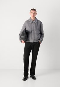 Gray herringbone jacket with a collar, zip closure, and two front pockets, paired with black trousers and leather shoes, holding a black bag.