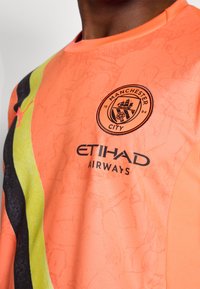 Narančasta Manchester City majica s crnim i žutim dijagonalnim prugama, s logom kluba i natpisom "ETIHAD AIRWAYS" u crnoj boji. Mekana tekstura.