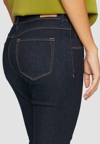 Dunkelblaue Denim-Jeans mit figurbetontem Design, kontrastierenden goldenen Nähten und zwei Gesäßtaschen. Etikett sichtbar am Bund.
