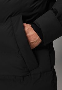 Chaqueta de plumón negra con textura acolchada, que cuenta con un bolsillo lateral. El puño está hecho de material acanalado negro, creando un ajuste ceñido.