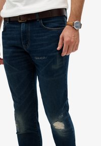 Jeans de denim azul oscuro con un corte ajustado, que presentan parches desgastados y desvanecimiento; combinados con un cinturón de cuero marrón y un reloj de pulsera.