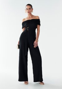 Black off-the-shoulder jumpsuit with textured fabric, wide legs, cinched waist featuring a gold ring belt, and accessorized with a small clutch.