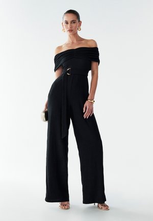 Black off-the-shoulder jumpsuit with textured fabric, wide legs, cinched waist featuring a gold ring belt, and accessorized with a small clutch.
