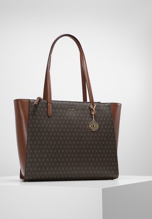 Brązowa torba typu tote DKNY z wzorem monogramu, gładkimi skórzanymi bokami, podwójnymi uchwytami oraz okrągłą złotą zawieszką z logo wiszącą u góry.