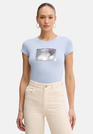 Donna che indossa una t-shirt aderente azzurro chiaro con toppa rettangolare di paillettes argento lucido, pantaloni beige a vita alta e orecchini a cerchio su sfondo uniforme.