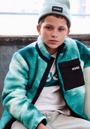 Jachetă din fleece turcoaz cu accente negre și buzunar frontal, purtată peste un tricou alb, asortată cu o căciulă albastru deschis.