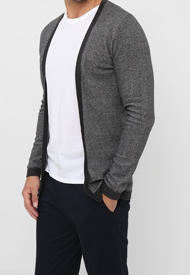 Grå stickad cardigan med ett strukturerat mönster, svart kant längs framkanterna, öppen frontdesign och långa ärmar. Bärs över en vit skjorta.