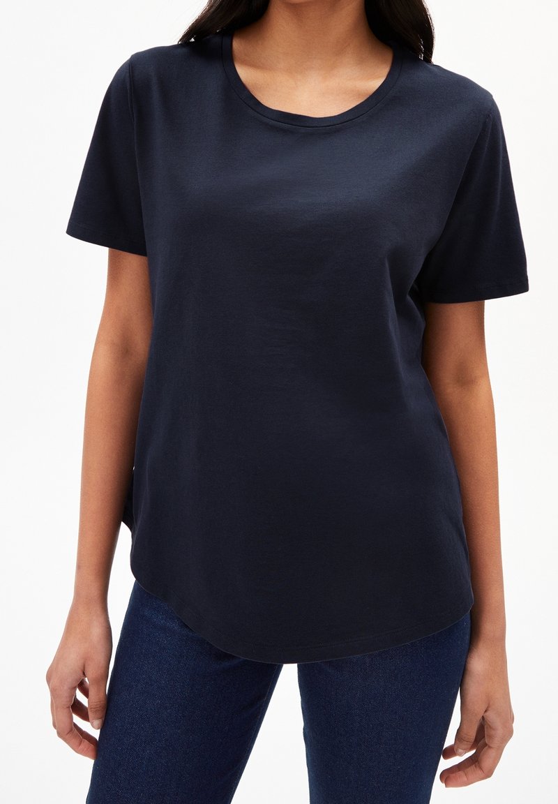Femme portant un t-shirt uni bleu marine foncé à manches courtes et un jean bleu foncé, debout devant un fond blanc.