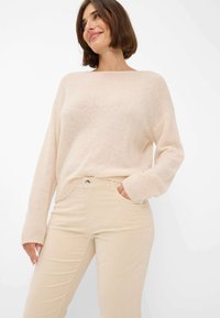 Lächelnde Frau in einem hellbeigen, gestrickten Pullover mit langen Ärmeln und palä beige Hochbundhosen, mit einer Hand in der Tasche, vor weißem Hintergrund.