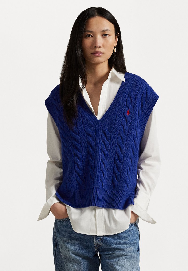 Blue cable-knit vest with a V-neck and ribbed hem, worn over a white button-up shirt. Features a red logo on the left chest area.