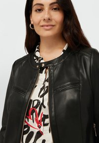 Black leather jacket with a round neckline, zip front, and decorative stitching on the shoulders and pockets, worn over a patterned shirt.