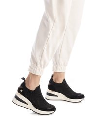 Zapatillas de punto negras con una superficie texturizada, media suela blanca y detalles dorados en el talón. Combinadas con pantalones de chándal blancos con puños.