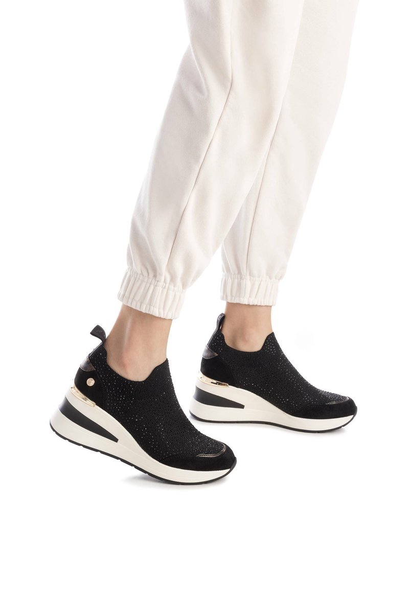 Zapatillas de punto negras con una superficie texturizada, media suela blanca y detalles dorados en el talón. Combinadas con pantalones de chándal blancos con puños.