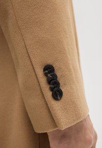 Close-up van een camel-kleurige mouw van een jas met vier zwarte knopen genaaid nabij de manchet, gedragen op de arm van een persoon.