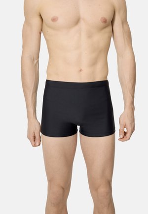 Torso masculino con bañador negro liso, brazos relajados a los lados, de pie contra un fondo blanco.