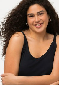 Top nero a costine con scollatura a V e spalline larghe, realizzato in un tessuto morbido e testurizzato. Il modello ha capelli ricci e incrocia le braccia.