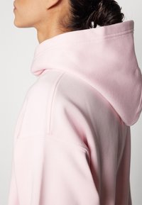 Felpa rosa chiaro con cappuccio ampio, texture morbida e dettagli di cucitura visibili sulle spalle e sul collo.