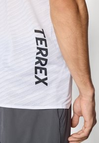 Right arm and hand beside white sports shirt with "TERREX" printed vertically, paired with gray pants.