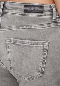 Los vaqueros de mezclilla gris presentan un bolsillo trasero con costuras visibles y una etiqueta negra que dice "ONLY". La tela tiene una textura deslavada.