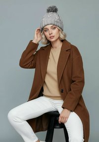 Brown wool coat over a beige sweater, paired with white pants. The model wears a gray knit beanie with a pom-pom, seated on a black stool.