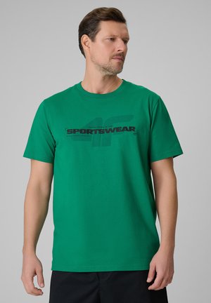 Mężczyzna w zielonej sportowej koszulce z krótkim rękawem z czarnym tekstem i wzorem, patrzący na bok na jednolitym szarym tle.