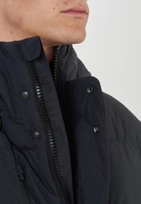 Giacca puffer blu navy con colletto alto, chiusura con zip, bottoni a pressione e dettaglio logo in rilievo. Tessuto morbido e testurizzato con finitura opaca.