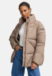 Beige pufferjack met een hoge kraag, gequiltde textuur, rits aan de voorkant en logo patch op de linkermouw. Gecombineerd met blauwe jeans.