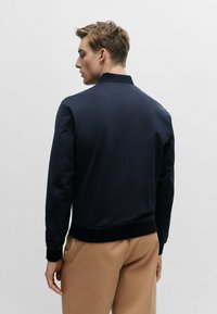 Navy bomber jacket with a smooth texture, ribbed collar and cuffs, and a fitted waist. Features minimal design with clean lines.
