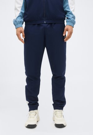 Homme portant un pantalon de survêtement bleu marine avec des poignets élastiques et des baskets couleur crème, debout devant un fond uni.