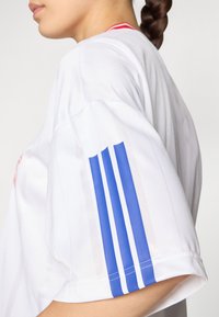 Witte sportshirt met een blauw driestroken ontwerp op de linkermouw en rode accenten dichtbij de kraag, met een soepele textuur.