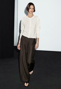 Una donna con un cardigan chiaro e pantaloni scuri cammina con sicurezza su un pavimento nero, con uno sfondo minimalista bianco e nero.