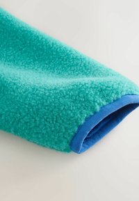Manica in pile turchese con superficie morbida e strutturata e bordatura blu. Il tessuto appare soffice e caldo, mettendo in risalto i colori a contrasto.