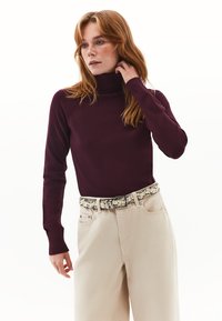 Bordeaux turtlenecktrui met lange mouwen, gemaakt van een zachte stof. Geverfd met crèmekleurige broek en een gestreepte riem.