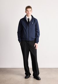 Μπουφάν αντιανεμικό σε ναυτικό μπλε χρώμα με φερμουάρ μπροστά, ελαστικά τελειώματα στα μανίκια και λογότυπο CK στο στήθος, σε συνδυασμό με μαύρο παντελόνι φόρμας και παπούτσια.