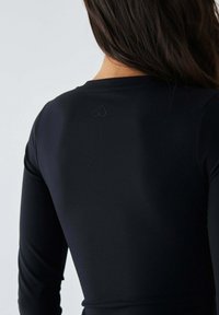 Schwarzes Langarmoberteil aus elastischem Stoff; verfügt über ein kleines, auf dem oberen Rücken eingeprägtes Herzlogo. Glatte Textur, körperbetonte Passform.