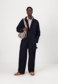 Ναυτικό μπλε φόρεμα με μακριά μανίκια, τσέπη στο στήθος με φερμουάρ και φαρδιά πόδια. Φορεμένο πάνω από πουκάμισο με καρό σχέδιο. Χακί παπούτσια και γκρι τσάντα ώμου.