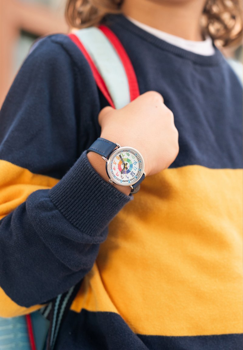Montre avec bracelet en cuir bleu, boîtier rond en acier argenté et cadran coloré affichant des chiffres dans un design adapté aux enfants, portée par-dessus un pull rayé bleu marine et jaune.